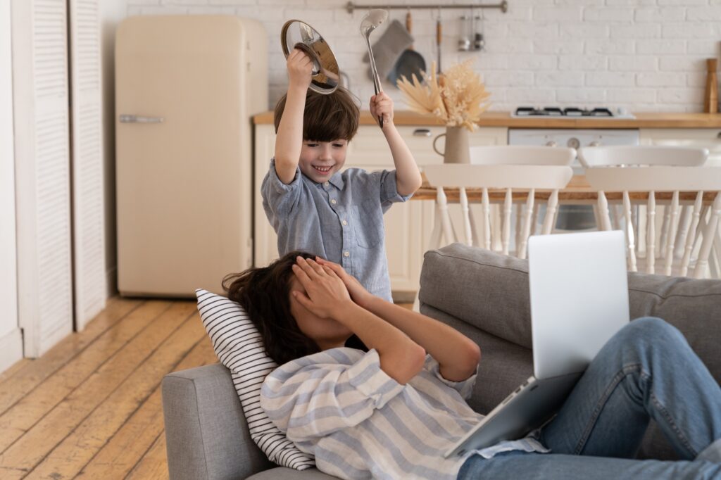 Unhappy young mom cover face with hand from noisy small son bad behavior. Mommy suffer from headache